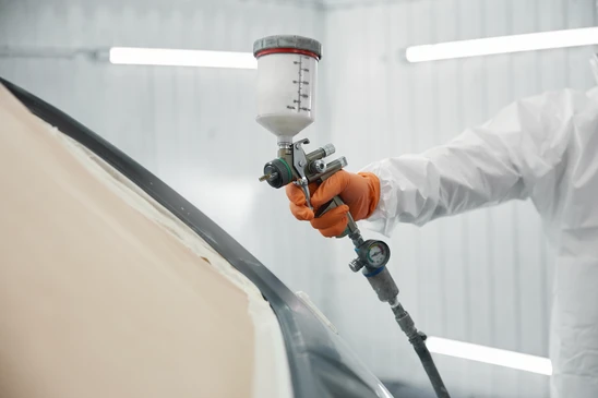 Cabine de peinture automobile professionnelle avec ouvrier en combinaison de protection intégrale appliquant de la peinture rouge au pistolet sur véhicule