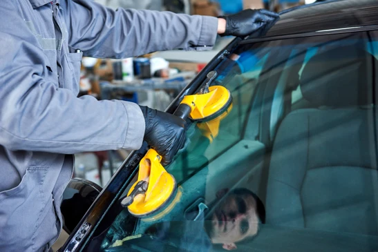 Atelier automobile intérieur, technicien en combinaison grise installant un pare-brise sur véhicule blanc à l'aide de ventouses industrielles bleues